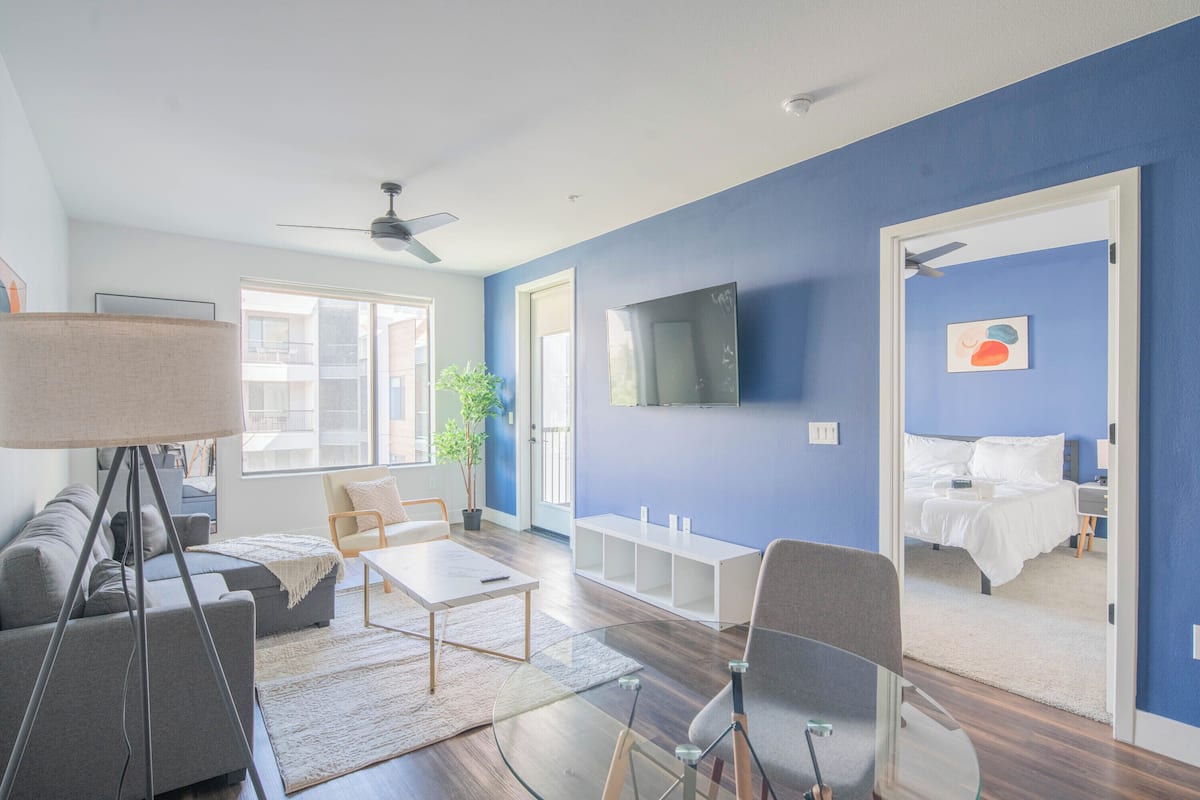 A modern living room with a sofa, coffee table, and chairs, a TV mounted on the wall, and a bed in the adjacent room.