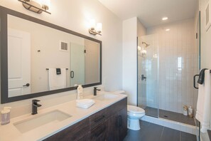 A modern bathroom with a large mirror, two sinks, a toilet, and a glass-enclosed shower area.