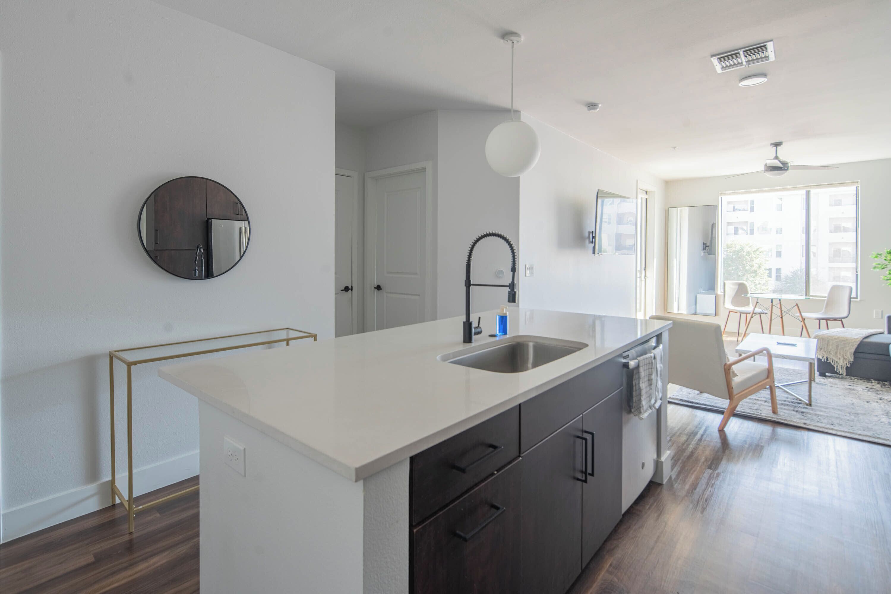 A modern kitchen with a white countertop, dark cabinets, and a round mirror. There is a dining area with a table and chairs visible through an open door.