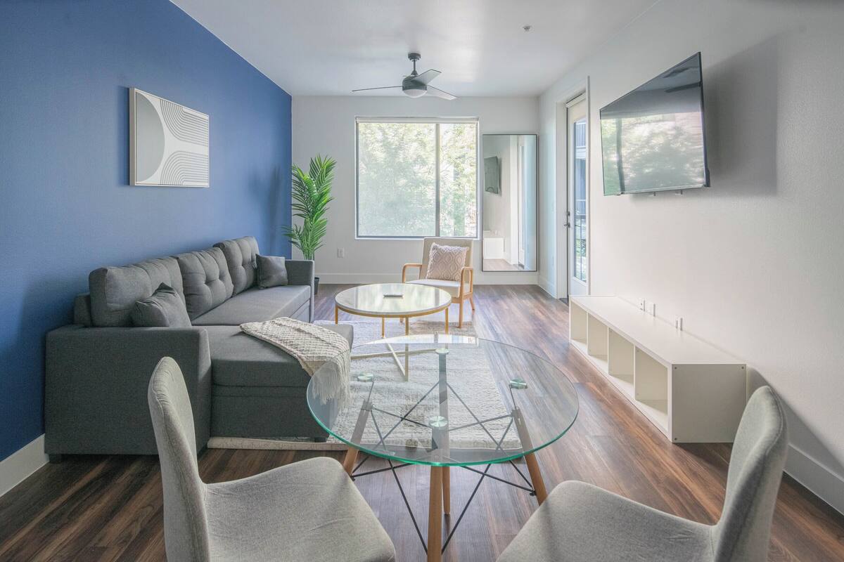 A modern living room with a glass coffee table, a grey sofa, and a large window.