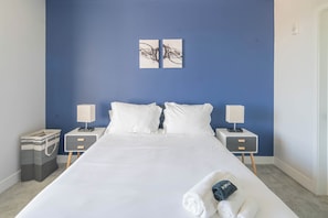 A neatly made bed in a room with blue walls, two bedside tables with lamps, and a pair of abstract wall art pieces.
