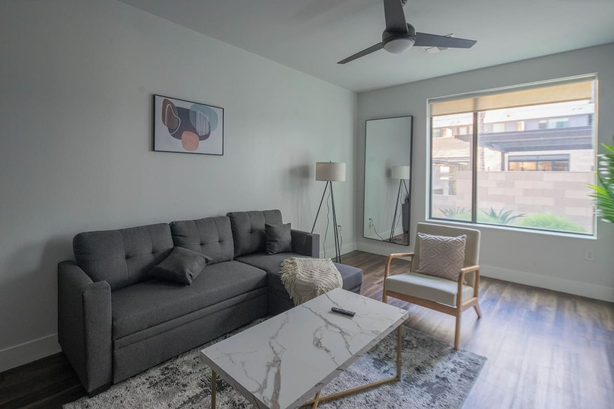 A modern living room with a grey sectional sofa, a white marble coffee table, a floor lamp, and a large window offering a view of the outside.