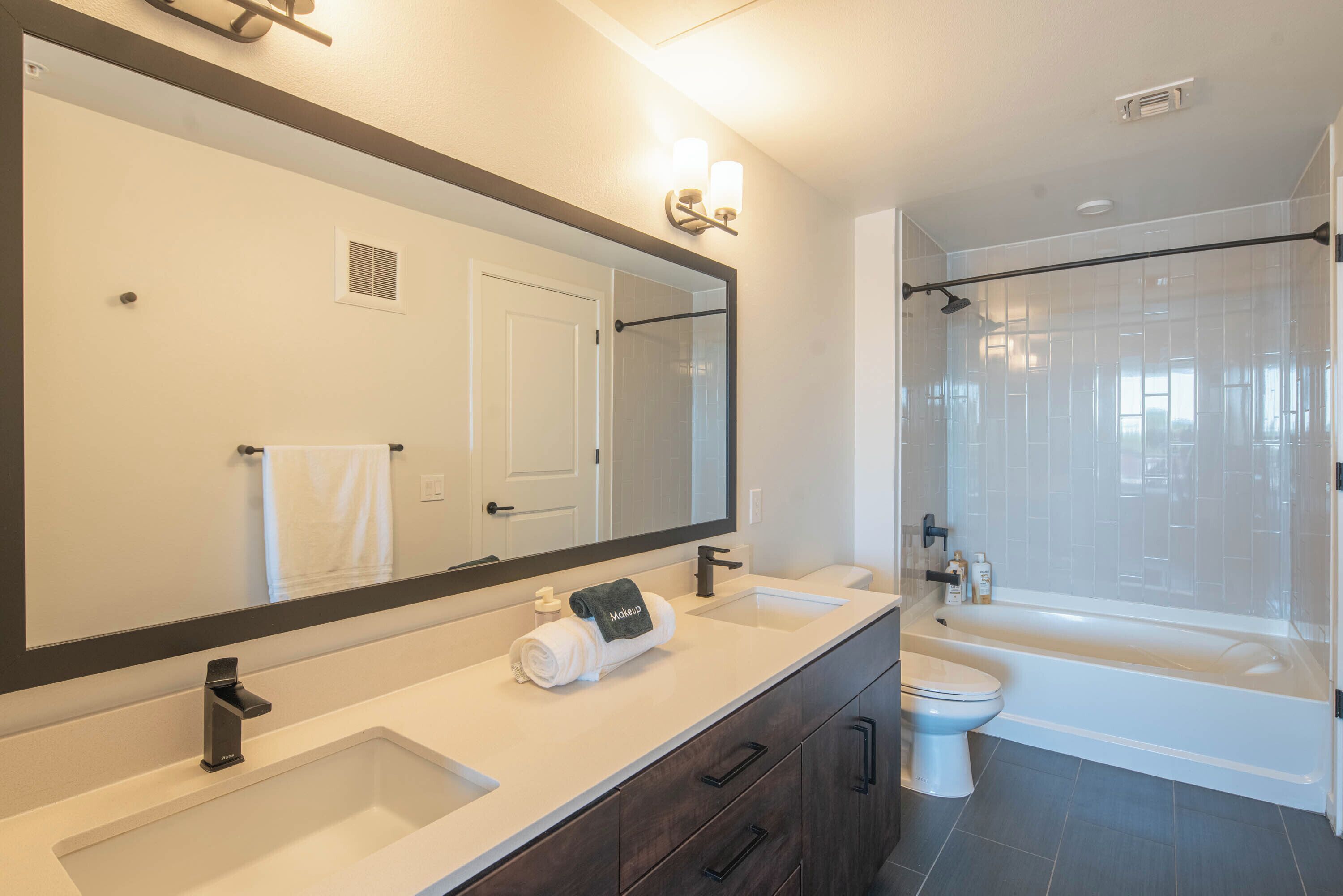 A modern bathroom with a large mirror, two sinks, a toilet, and a bathtub.