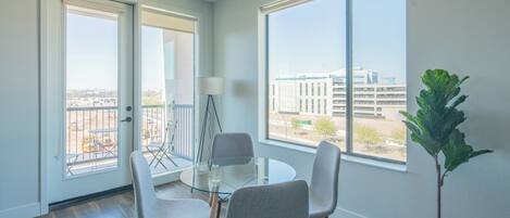 Una habitación moderna con una mesa de vidrio y cuatro sillas, ventanas amplias con vistas a edificios, y una planta en maceta.