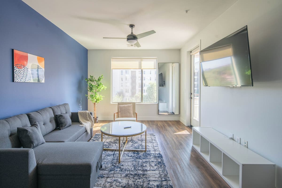 A modern living room with a grey sofa, a round coffee table, a ceiling fan, a large window, and a TV mounted on the wall.