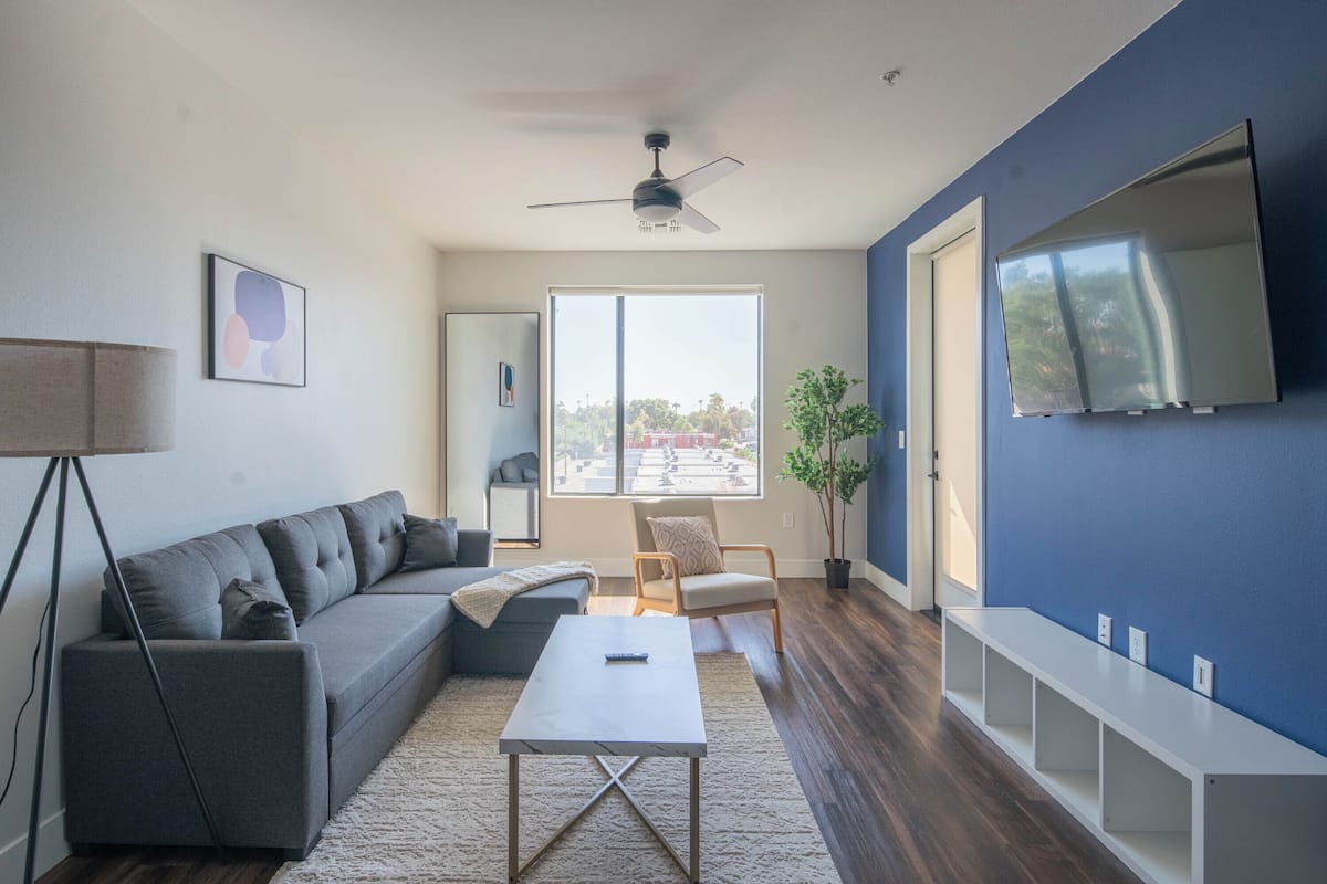 A modern living room with a grey sofa, a white coffee table, a ceiling fan, a large window, and a TV mounted on the wall.