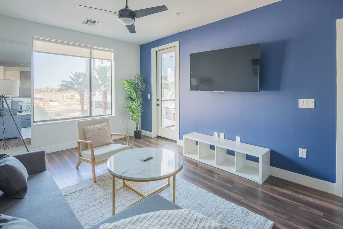 A modern living room with a sofa, a round coffee table, a TV mounted on the wall, and a large window with a view of palm trees.