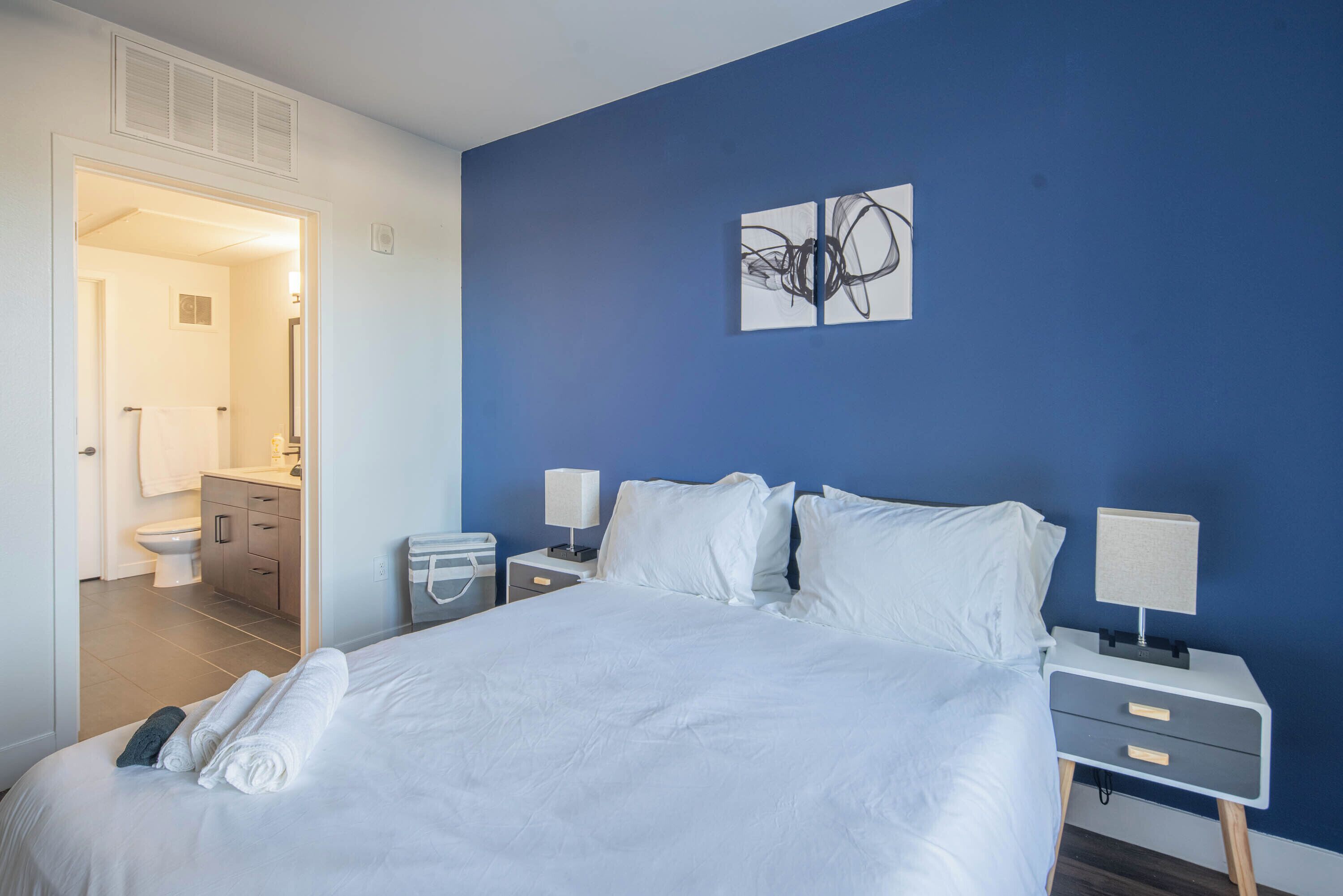 A hotel room with a large bed, two bedside tables, a nightstand, a bathroom visible through an open door, and a blue accent wall.