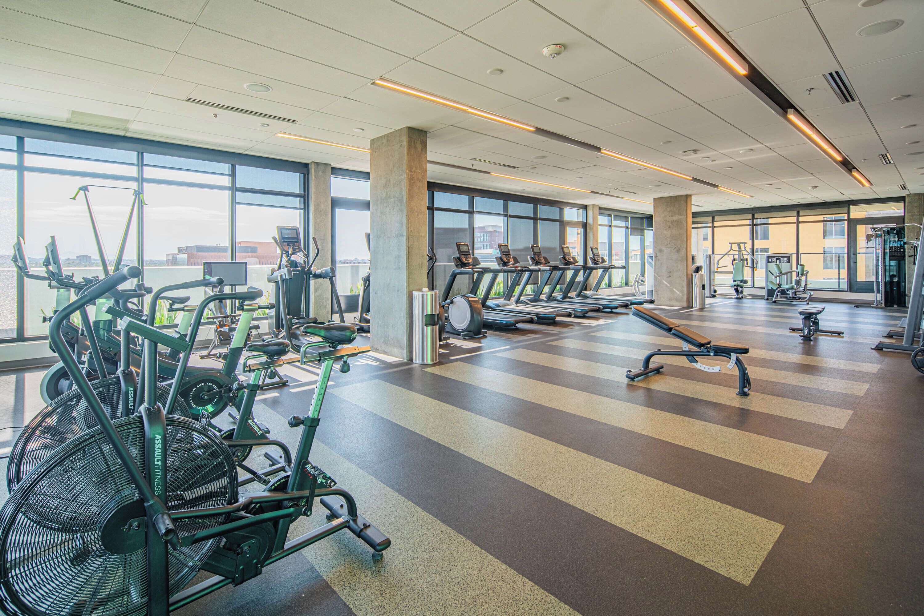 A spacious gym with various exercise equipment, large windows, and a high ceiling.