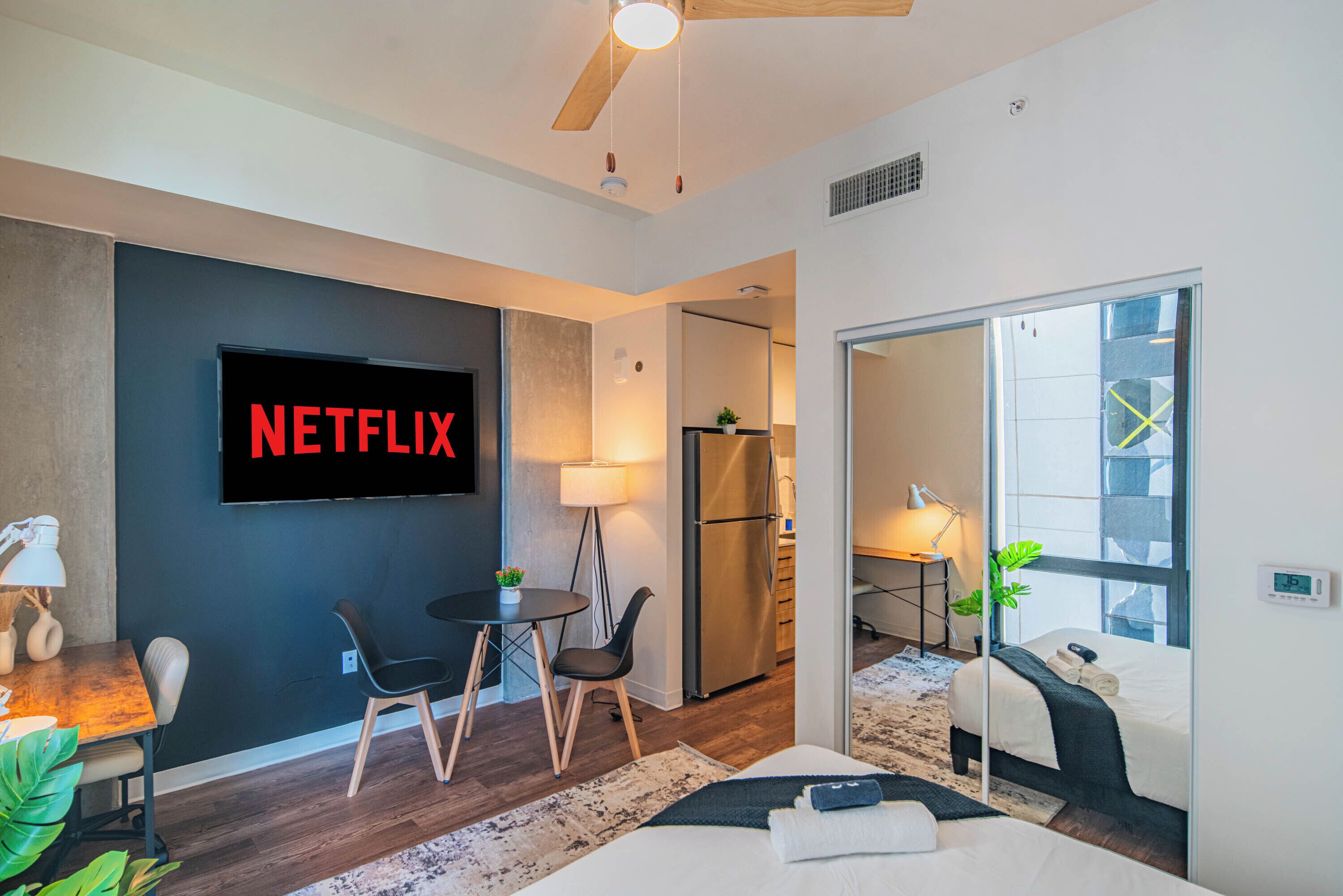 Une chambre d’hôtel moderne dotée d’un grand téléviseur à écran plat affichant « NETFLIX », d’un coin repas avec une table et des chaises, d’un lit avec un miroir et d’une vue sur l’extérieur grâce à une porte vitrée.