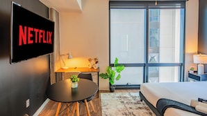A modern hotel room with a large flat-screen TV displaying the Netflix logo, a comfortable bed, a small desk with a lamp, a potted plant, and a large window offering a city view. - ROVE LIVING/KB - Joy on 4th (Phoenix)