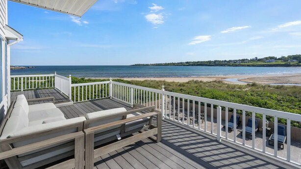 Eine Holzterrasse mit weißen Geländern, Sitzmöbeln im Freien und Blick auf Strand und Meer.