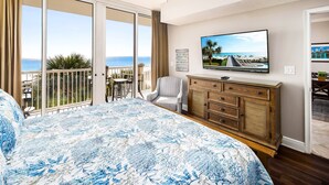 A bedroom with a large bed, a TV, a wooden dresser, and a balcony with a view of the ocean. - Waters Edge 304 • Luxury 3BR 3BA, Balcony, Large Pool, Covered Parking, Free Bch Service (Fort Walton Beach)