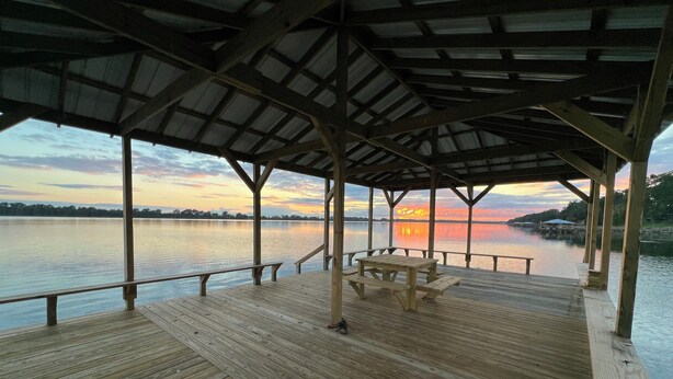 Eine Holzterrasse mit überdachtem Bereich und einer Bank, mit Blick auf ruhiges Wasser bei Sonnenuntergang.
