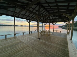 A wooden deck with a covered area and a bench, overlooking a calm body of water during sunset.