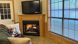 Ein Wohnzimmer mit Kamin, Fernseher auf einem Schrank, einer Couch und einem großen Fenster mit Jalousien.