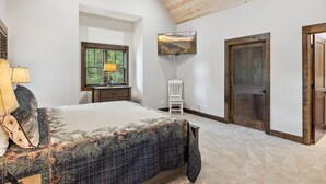 A bedroom with a large bed, a window with a view of greenery, a white chair, and a wooden door.