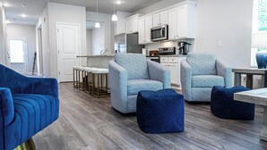 Una cocina moderna con armarios blancos, microondas y refrigerador de acero inoxidable. En la sala de estar, hay dos sillones azules y un otoman azul.