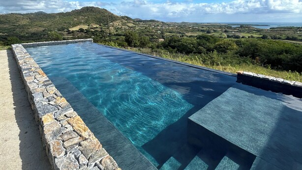 A clear blue infinity pool with a stone border, surrounded by a scenic landscape with hills and a clear sky. - Ecolodges Puyferrat (Le Marin)