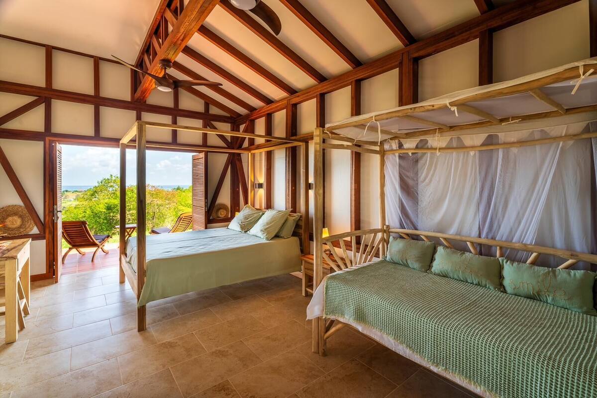 A room with a bed, a chair, and a table. There is a view of greenery outside.