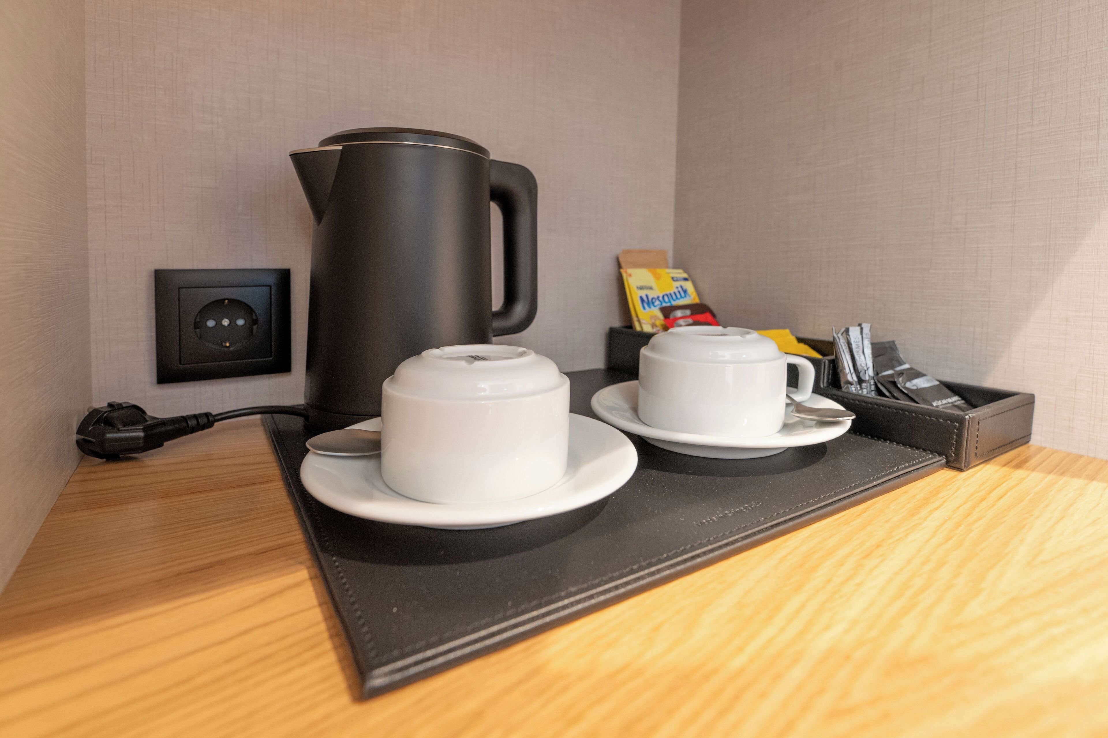 Une chambre d’hôtel avec une machine à café noire, deux tasses blanches sur un plateau noir, une prise électrique et une boîte de Nesquik.