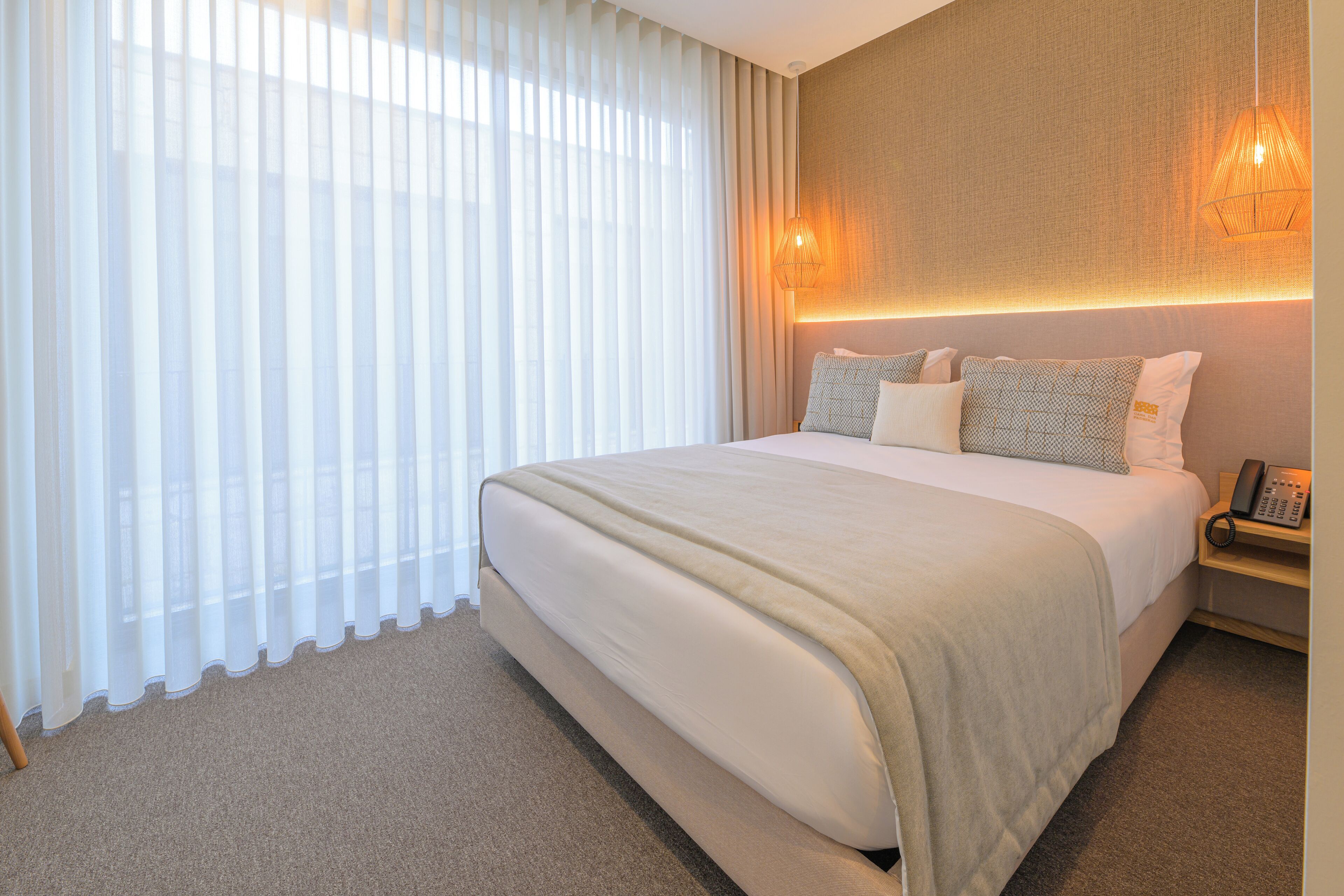 A hotel room with a large bed, a bedside table with a lamp, and sheer curtains on the window.