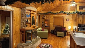 A cozy cabin interior with a stone fireplace, wooden walls, a green sofa, and a small kitchen area.