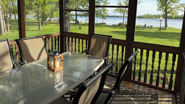 A covered outdoor dining area with a glass table and chairs, overlooking a grassy area and a lake.