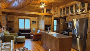 A cabin interior with a wooden interior, a kitchen with a stainless steel refrigerator, and a seating area with a sofa and armchairs.