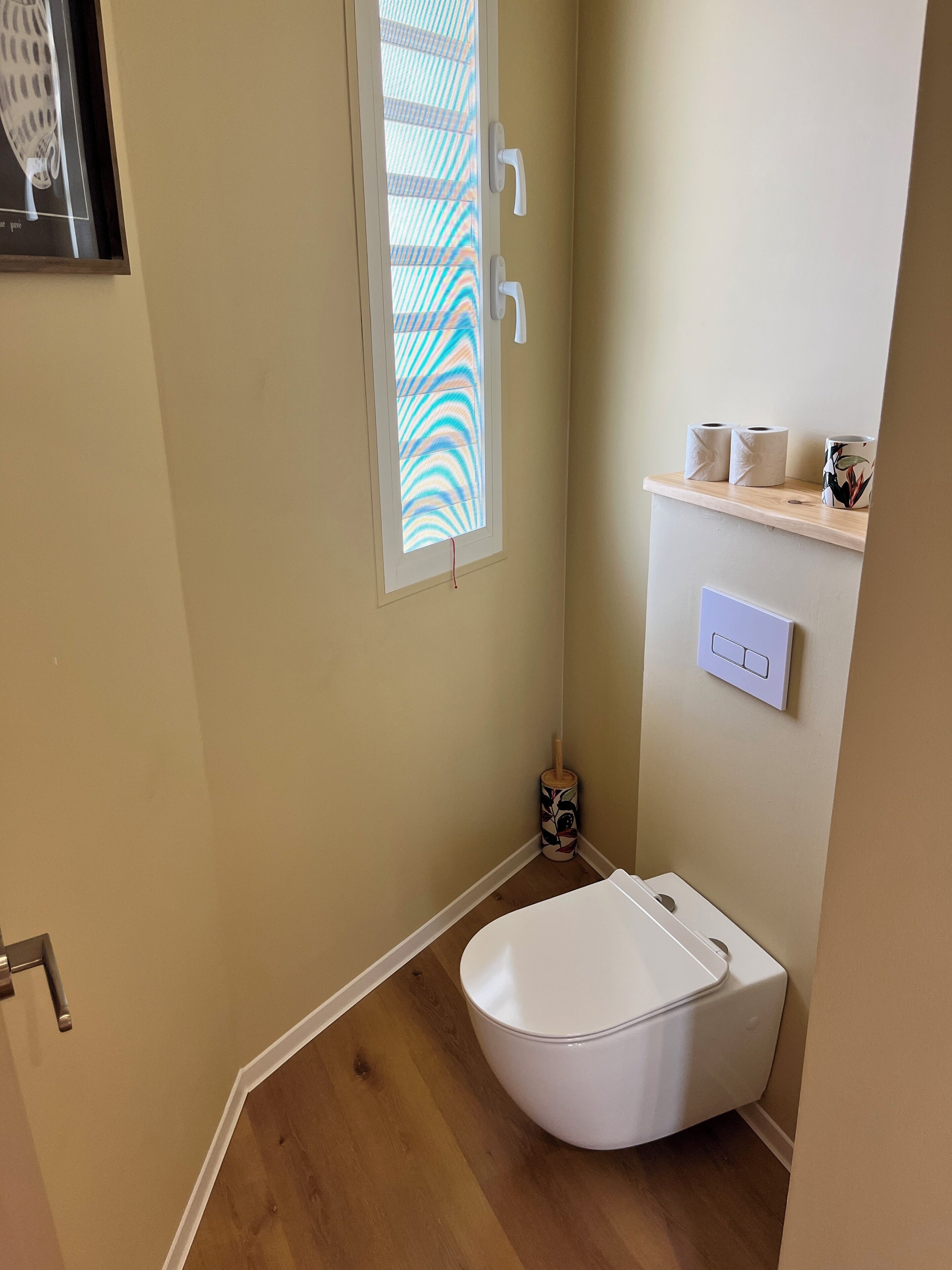A small bathroom with a white toilet, a window with a striped pattern, a toilet paper holder, and a wall-mounted light switch.