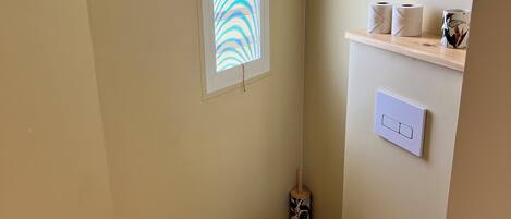 A small bathroom with a white toilet, a window with a striped pattern, a toilet paper holder, and a wall-mounted light switch.