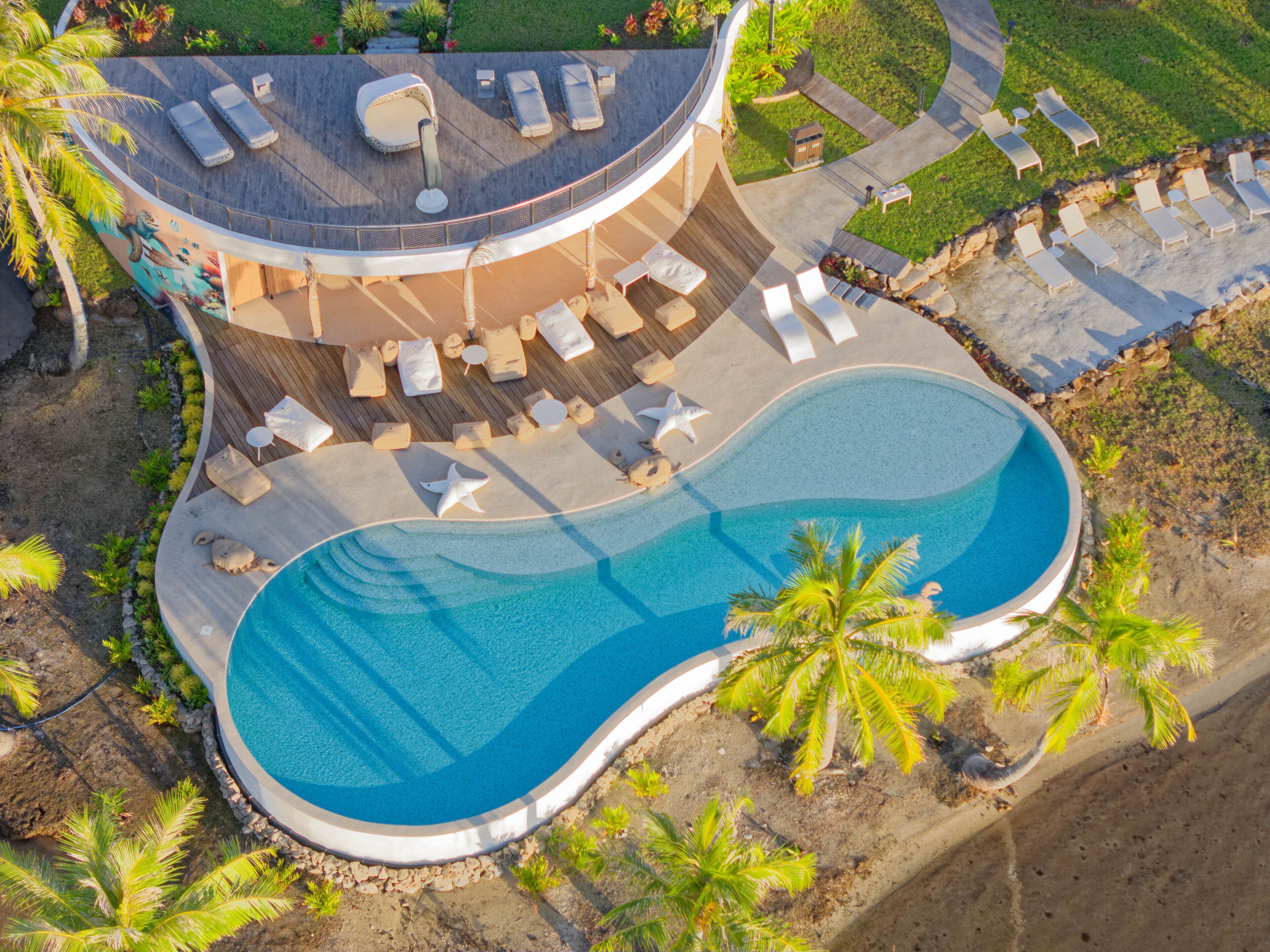 Luftaufnahme eines Resorts mit großem Pool, Liegestühlen und Palmen.