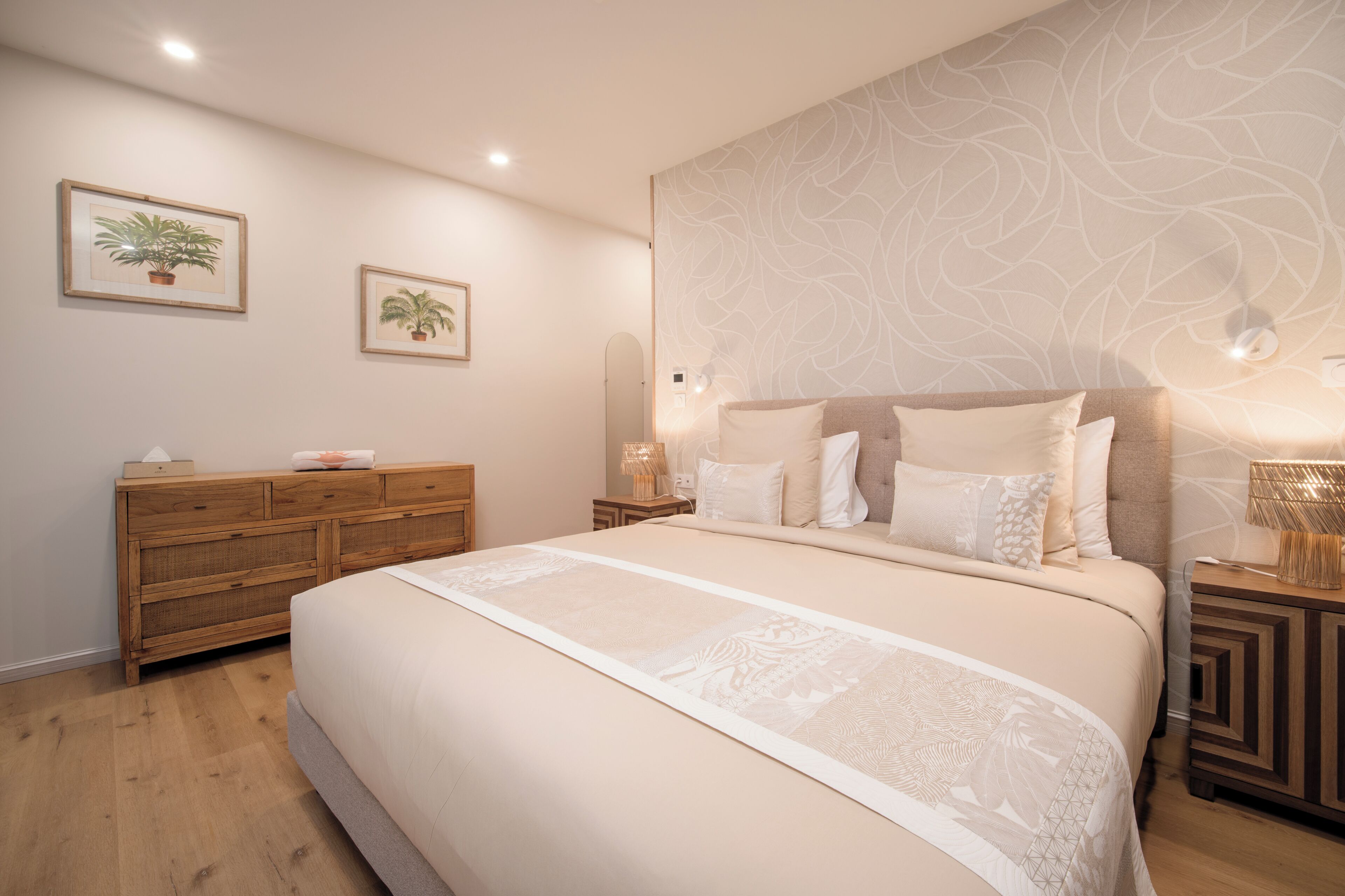 Un dormitorio con una cama grande, cómoda de madera, dos plantas enmarcadas y un papel tapiz estampado.