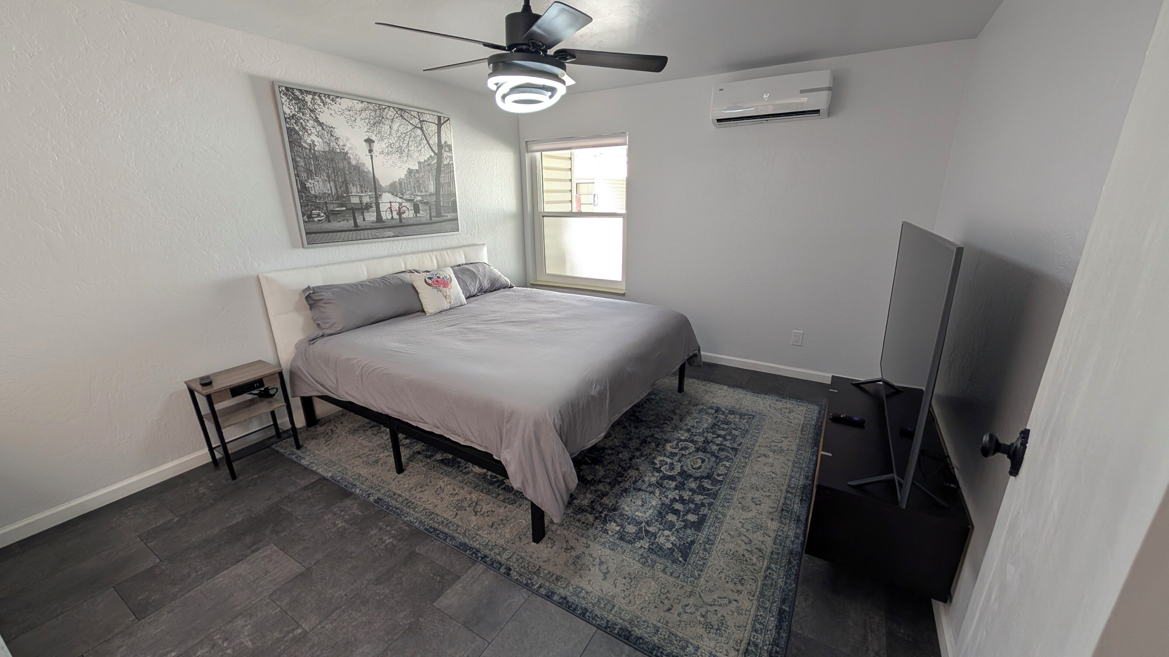 A bedroom with a bed, a ceiling fan, an air conditioner, a window, and a TV.