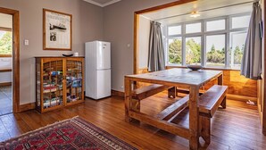 Un comedor con una mesa de madera y bancos, un refrigerador blanco, un armario de vidrio con libros y un ventanal con cortinas.