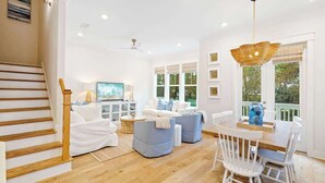 A spacious living area with a staircase, a television, a dining table with chairs, and a large window with a view. - OnHoliday30A by Stay on 30a (Inlet Beach)
