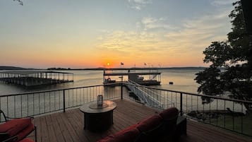 Une terrasse en bois avec une table ronde et des sièges rembourrés de couleur rouge, donnant sur une étendue d’eau calme et un quai orné de drapeaux.