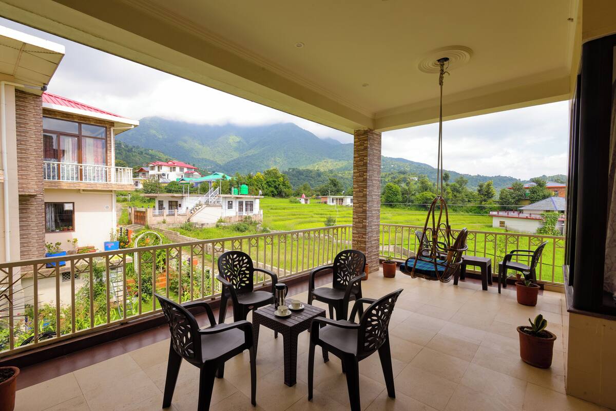 Ein Balkon mit Blick auf eine grüne Landschaft und Berge. Vier Stühle und ein Tisch mit Wasserkocher und Tassen stehen bereit.