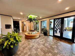 A hotel lobby with a central seating area, a reception desk, and a view of the dining area through glass doors.