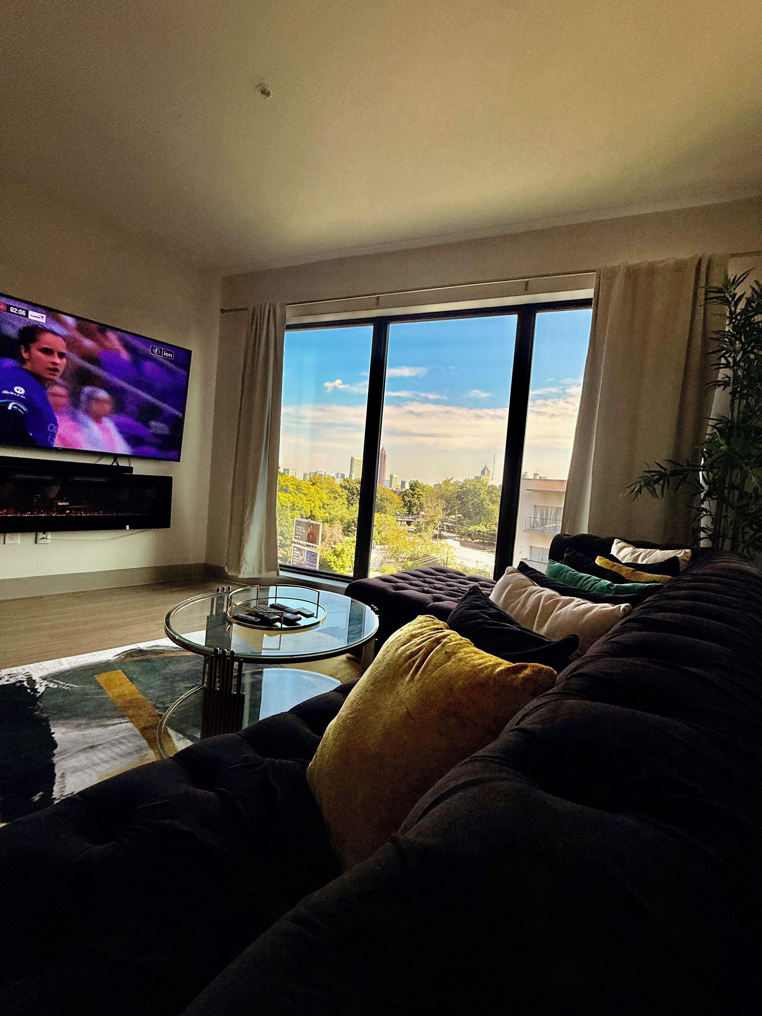 A living room with a large flat-screen TV mounted on the wall, a glass coffee table, a dark-colored sofa with multiple cushions, and a large window offering a view of a cityscape.