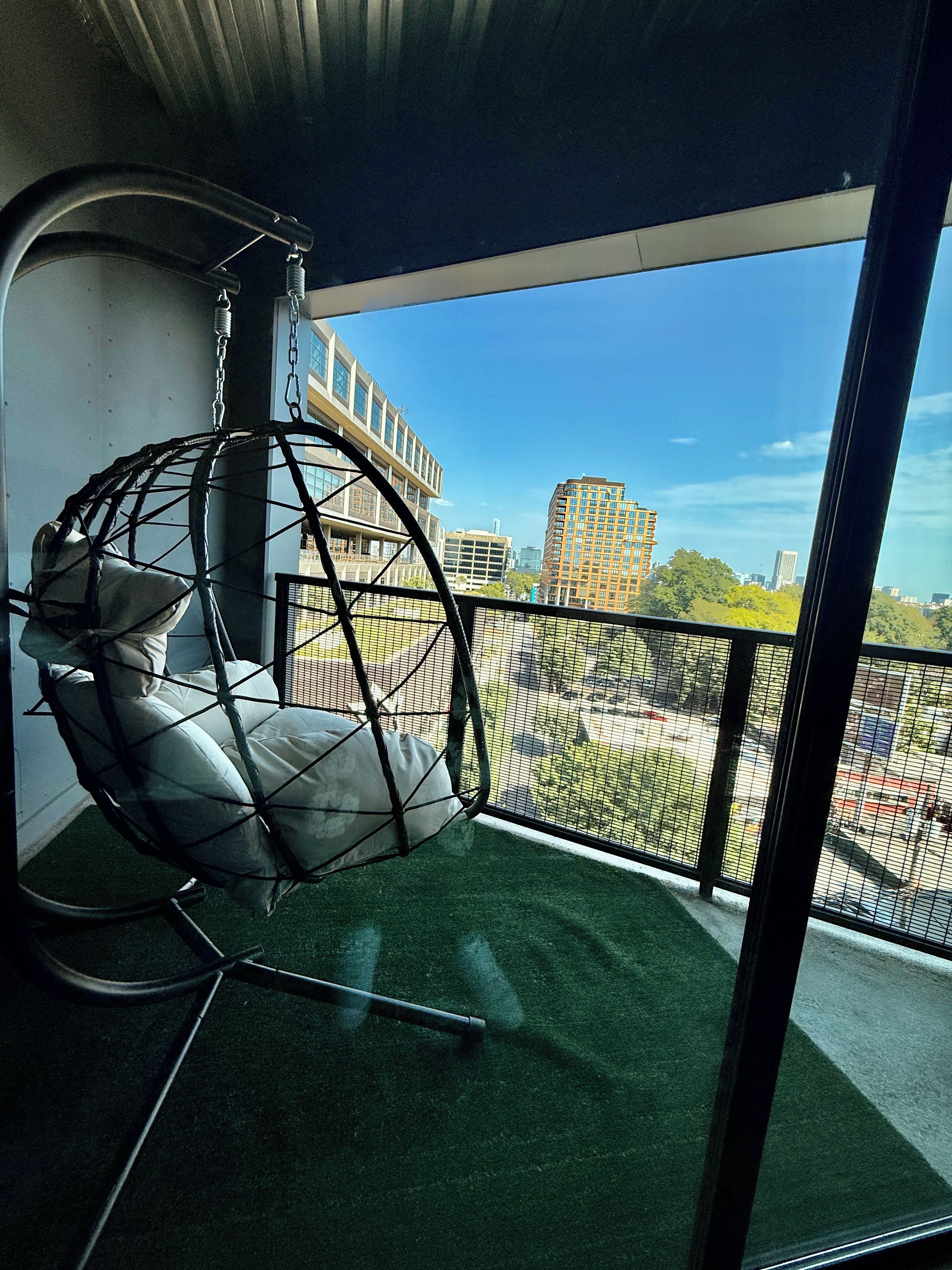 Una habitación con una silla colgante, vistas de un paisaje urbano a través de un ventanal grande y un balcón con barandilla.
