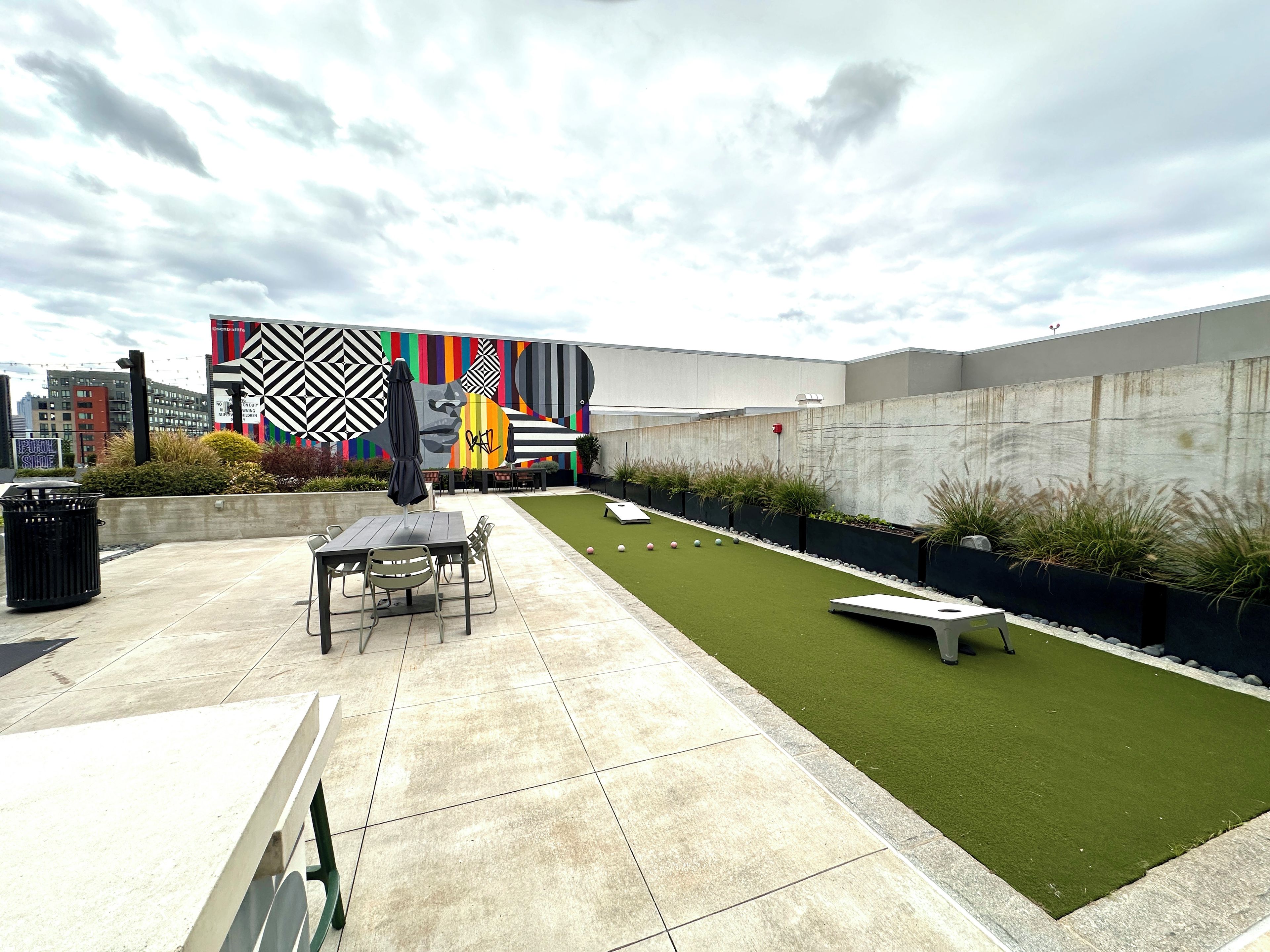 A rooftop patio with a dining table, chairs, a trash bin, and a small garden area with artificial grass and plants.