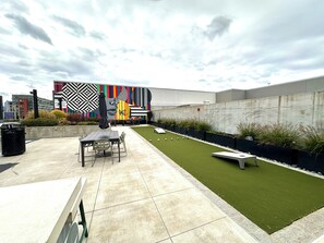 A rooftop patio with a dining table, chairs, a trash bin, and a small garden area with artificial grass and plants.