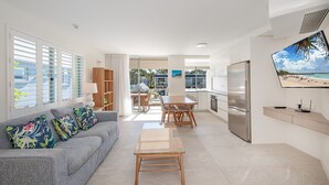 Una sala moderna con un sofá gris, una mesa de centro de madera y cojines coloridos. Hay un comedor con una mesa y sillas de madera, y una cocina con un refrigerador de acero inoxidable y un televisor de pantalla plana montado en la pared.