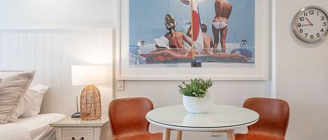Habitación de hotel con cama, mesita de noche, lámpara, cuadro enmarco, dos sillas, una pequeña mesa con una planta y un reloj de pared.
