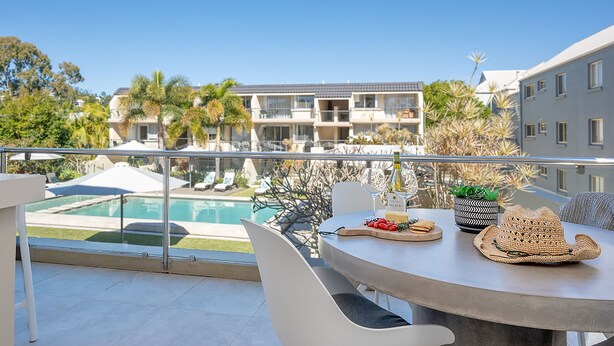 Un balcón con vista a la piscina, una mesa con un sombrero y una botella de vino.