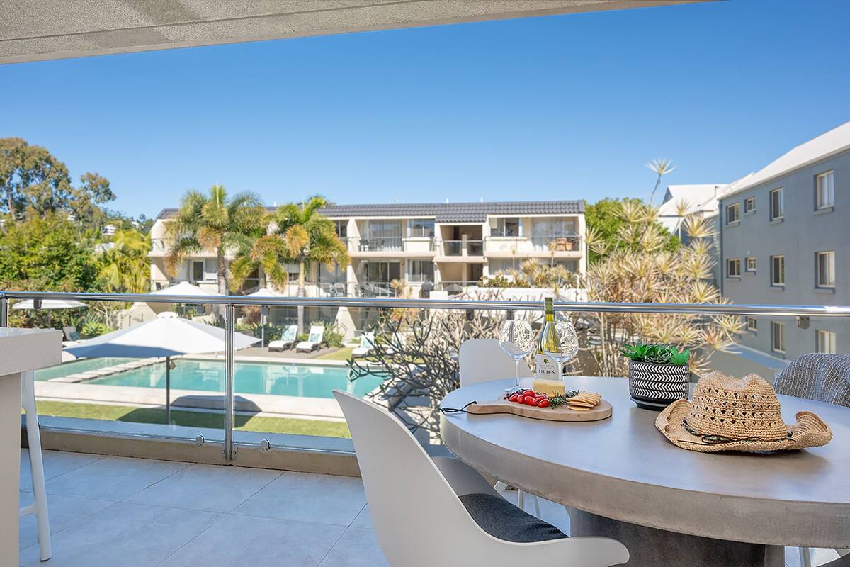 Un balcón con vista a la piscina, una mesa con un sombrero y una botella de vino.