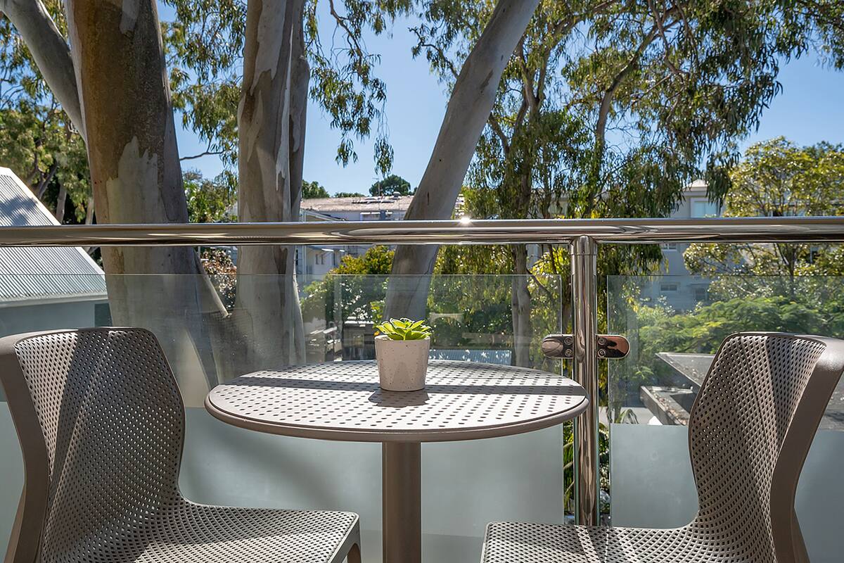 Un balcón con dos sillas de metal, una mesa redonda con planta en maceta y barandilla de vidrio con vistas a una zona residencial con árboles y edificios.