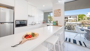 Una cocina moderna con armarios blancos, una isla grande con una tabla de frutas y quesos, y vista a una casa de playa a través de una puerta corrediza de vidrio.