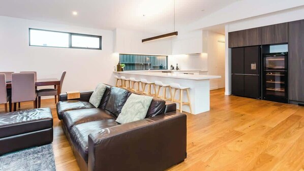 A modern living room with a leather sofa, a dining area with chairs, a kitchen with a bar, and a large window.